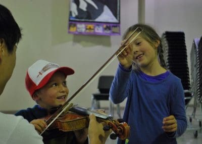 One child plays a violin while another looks at the bow at the CB Music Festival Sound Track Program for Kids.