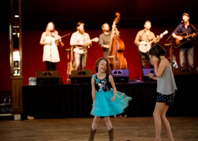 Two girls dance in front of a stage with musicians playing stringed instruments including a fiddle, mandolin, bass, banjo, and guitar at the CB Music Festival. Photo by Xavier Fane.