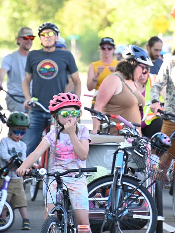 A community bike ride with familes, bikes, bike trailers, and all ages of people. A little girl with a pink helmet is in sharp detail in the foreground.