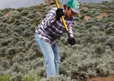 A member of the Gunnison Trails Youth Crew hacks out a path on a sagebrush hill with a pick axe.