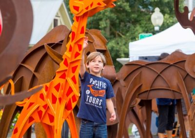 Arts Festival boy reaching up to touch an orange giraffe sculpture.