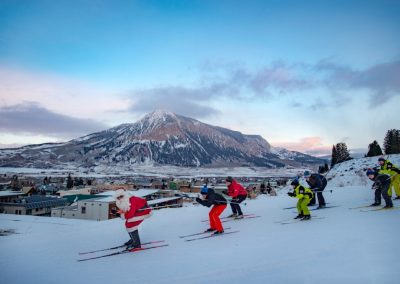 A group of skiers are going fast down a slope with Santa in the lead. Mt Crested Butte and the town of Crested Butte are in the backdrop. Photo by Xavier Fane.