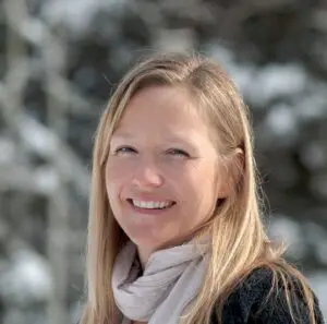 Jenny Birnie wearing a winter scarf and smiling in the sun against a snowy backdrop.