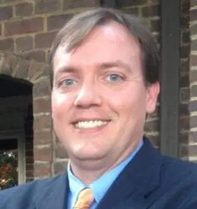 Daniel Bruce in a coat and tie in front of an interior brick wall.