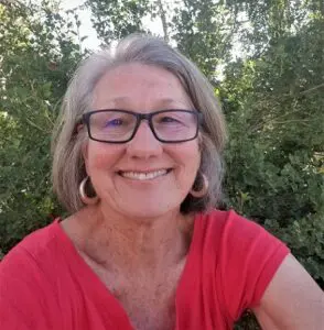 Patricia Harrington smiles in a summer scene with a red shirt and dark rimmed glasses.