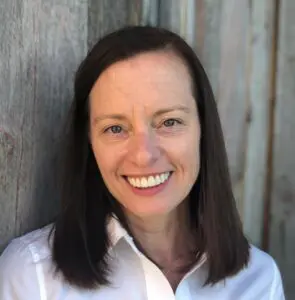 Paula Mann smiles in front of a gray wood wall in a white shirt.