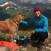 Tina McGuinness and her golden retriever smile from a mountain peak with a valley far below.