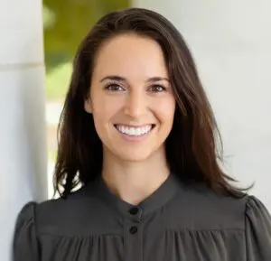 Lindsay Newman smiles wearing a black shirt with white columns and sunshine on green hedges behind her.