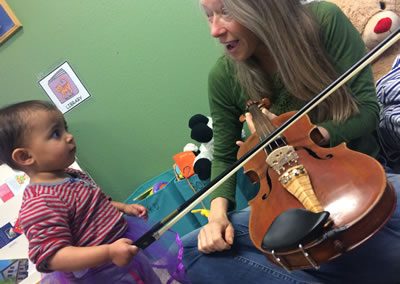 A teacher encourages a toddler to play a violin. Paradise Place School Endowment.