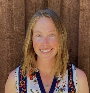 Brittany Perkins smiles with medium length blond hair and blue eyes wearing a floral blouse in front of a wooden wall.