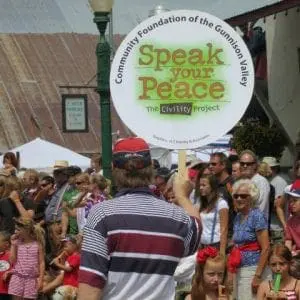 A Speak Your Peace Sign is held up in the Crested Butte Parade with lots of people watching in the crowd.