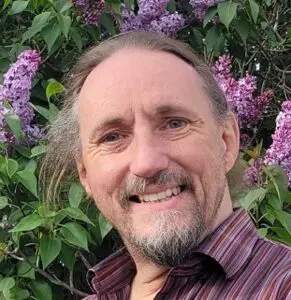 William Spicer smiles sporting a beard standing in front of blooming lilacs flowers.