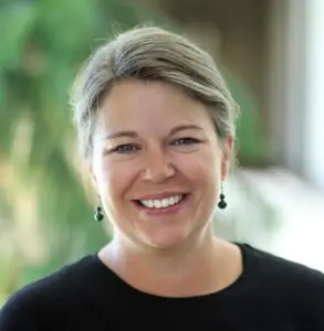 Kelly Sudderth smiles with her hair up wearing dangle earrings and a black top with a backdrop of greenery.