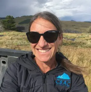 Karen Williams smiling wearing dark sunglasses in a breezy location with sagebrush and grassy hills.