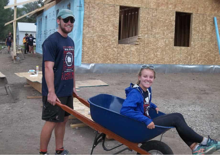 Two volunteers at a Habitat for Humanity build laughing while one hauls the other around in a wheelbarrow.