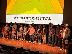 Crested Butte Film Festival participants line up on the stage under a big bnner of yellow, green, and orange freeform shapes.