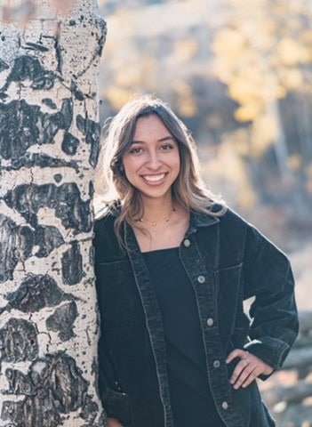 Greisy Martinez Davalos leaning against a knotty aspen tree with a smile