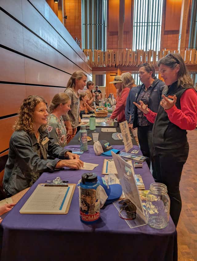 one woman sitting behind a table with information speaks to another standing and asking questions