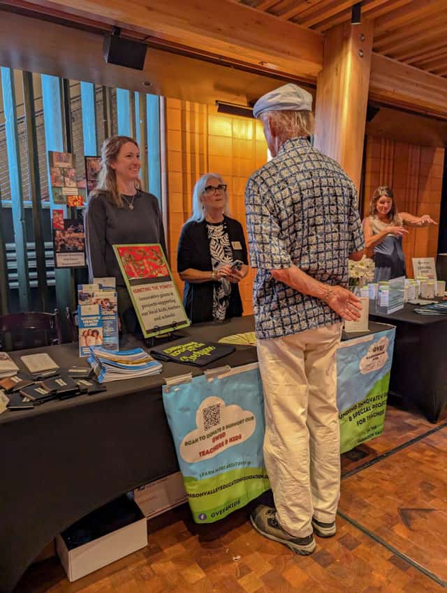 man with his back to the camera speaks to two women standing behind a table with material about the Gunnison Valley Education Foundation