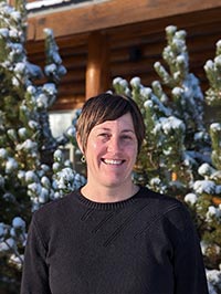 smiling woman with short hair wearing a black shirt in front of snowy trees and a log building