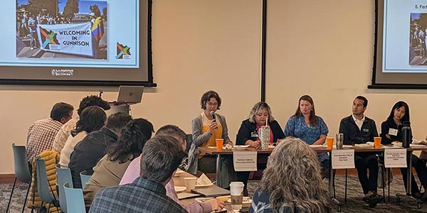 group of people sitting at table facing away from camera and looking at a group of panelists, one with a microphone, speaking about the issue of welcoming in Gunnison