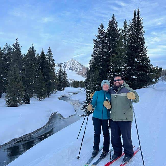 smiling couple on Nordic skis near a river with Crested Butte Mountain in the background