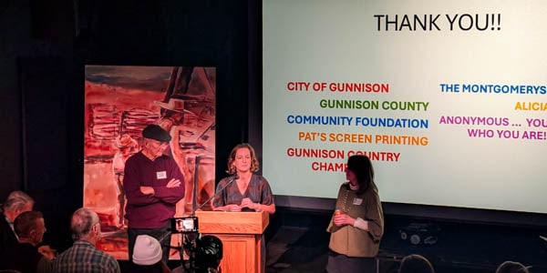 George Sibley, Amanda Cook, and Maryo Ewell standing behind a podium at the Gunnison Arts Center theater with a painting and a thank you message on a screen projected behind them.