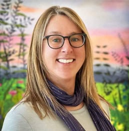 Carlie Kenton in front of a colorful mural of a mountain sunset wearing a mid-length hairstyle and purple scarf.