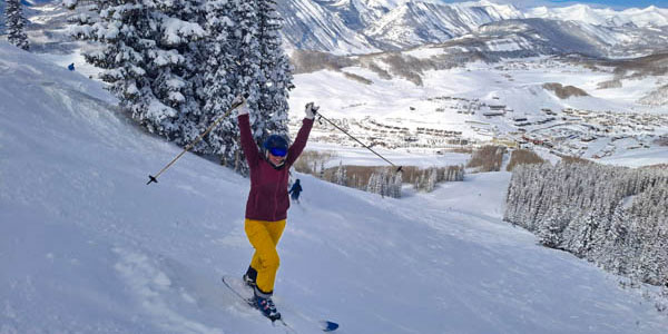 smiling woman wearing blue helmet, magenta jacket, yellow snowpants, and blue ski boots standing on skis on fresh snowy trail with mountains in the background