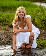 Dayla Smith young lady in white dress stoops down to put her hand in a brook