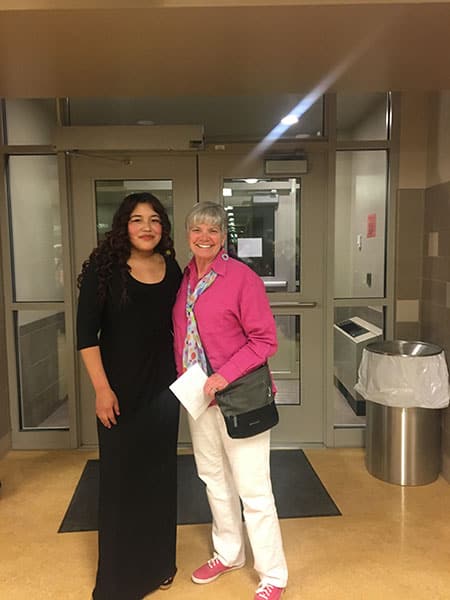 2025_Rita_and_Aida Two women standing together. The one on the left has long, curly black hair and is wearing all black and smiling. The one on the right has short silver hair and a big smile. She's wearing a pink jacket, white pants, and pink shoes.