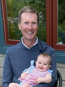 Smiling Bill wearing a dark gray shirt and holding a smiling baby wearing a red, white, and blue striped outfit