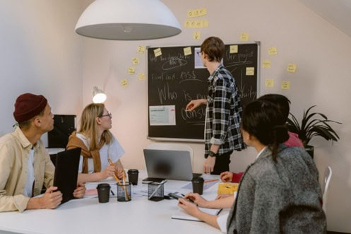 A group of people sit around a white table and look at a man in a flannel shirt point to something written on a black chalkboard that's surrounded by sticky notes